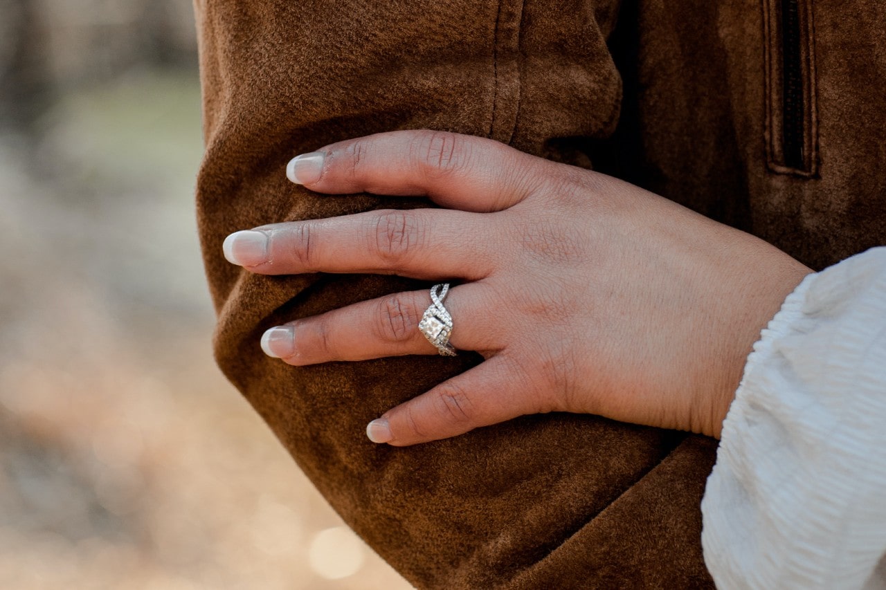 A diamond and moissanite ring on a woman&rsquo;s hand resting on the elbow of her partner wearing a tan suede jacket