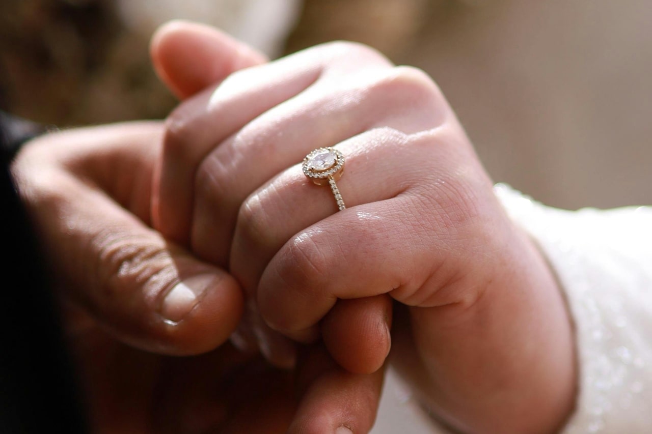 a man&rsquo;s hand holding a woman&rsquo;s that is adorned with an oval cut halo set engagement ring