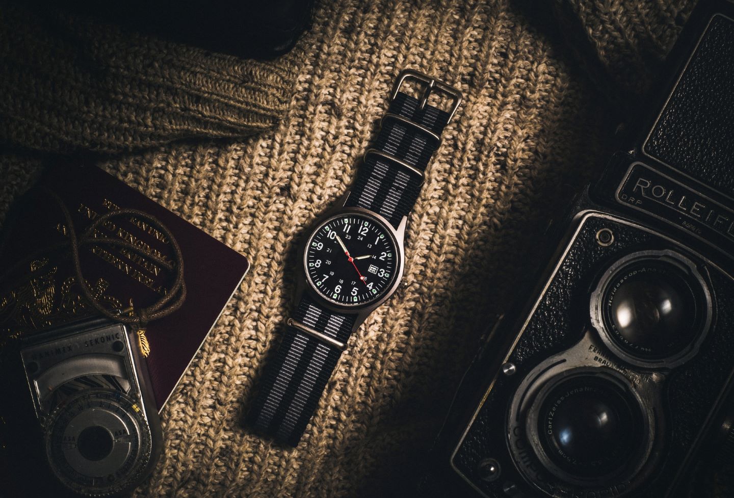 A watch with a nylon strap sitting next to a camera, atop a knit sweater.