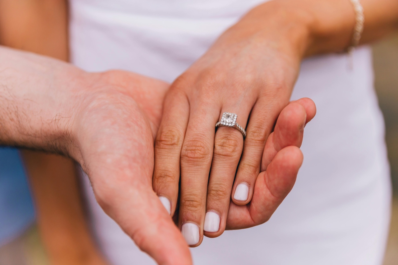 A close-up of a man’s hand holding a woman’s hand, adorned with a princess cut engagement ring.