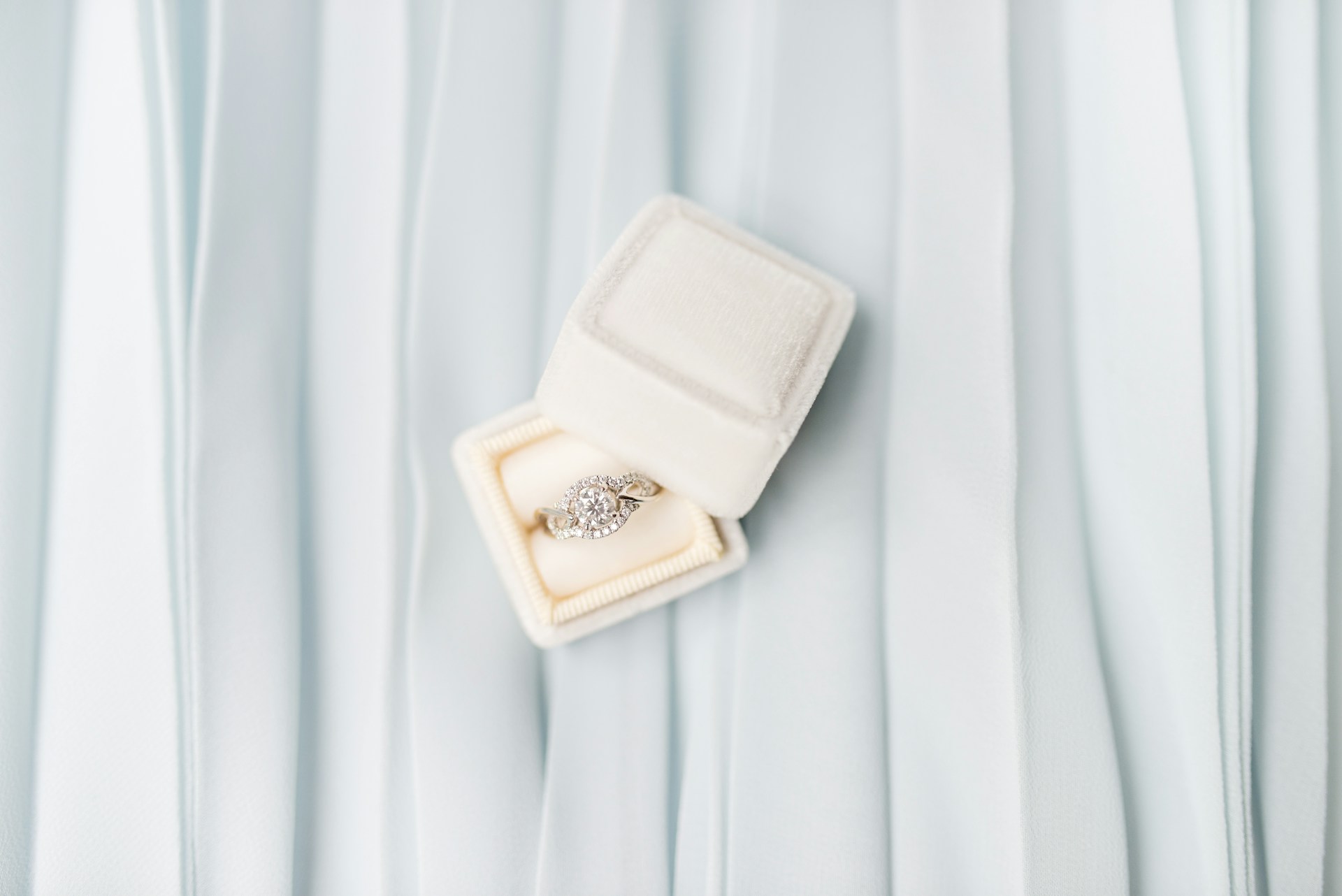 A halo engagement ring in a white ring box, sitting on a piece of blue fabric.