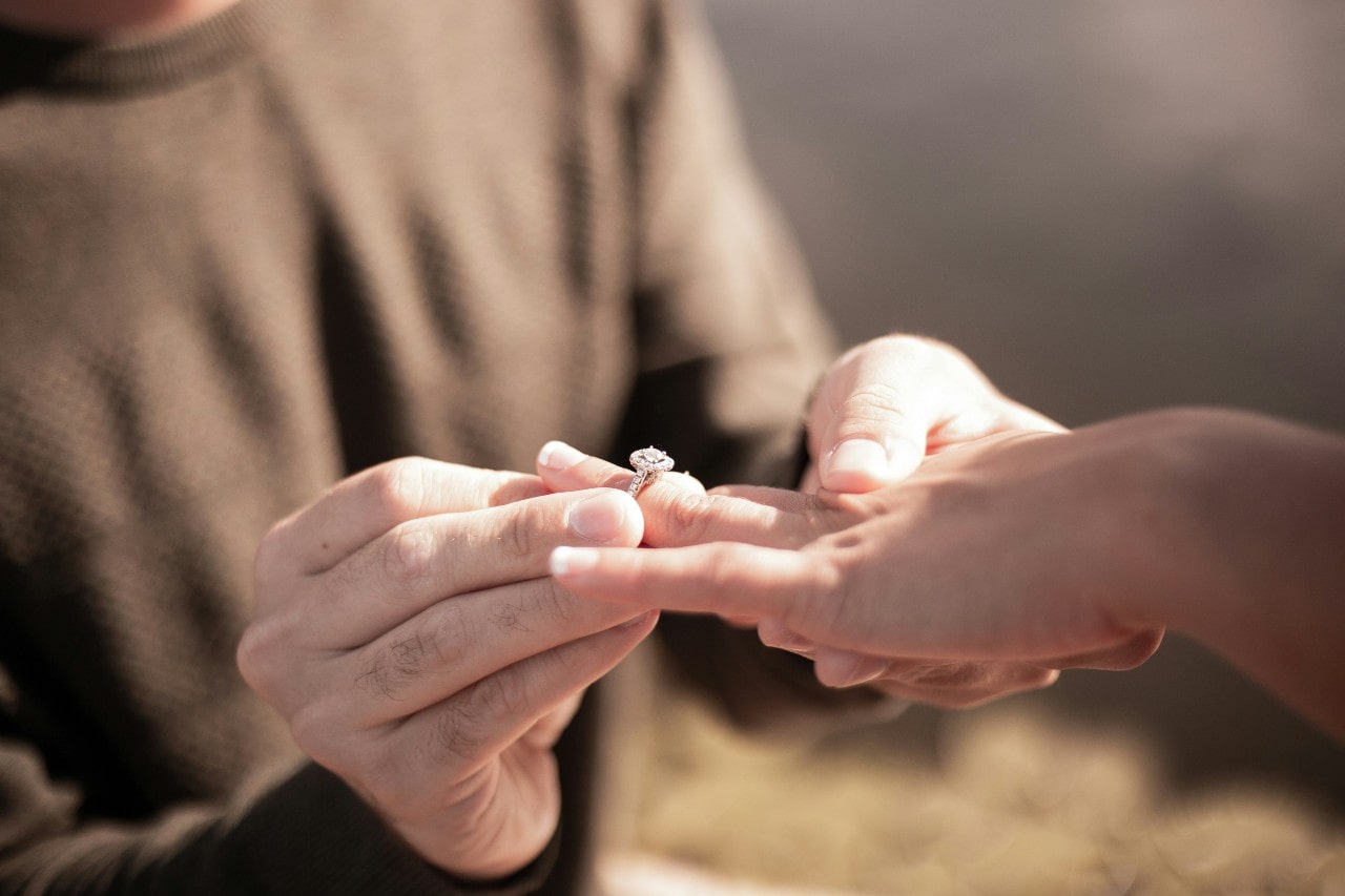 A close-up of a man placing a diamond engagement ring on a woman’s finger.